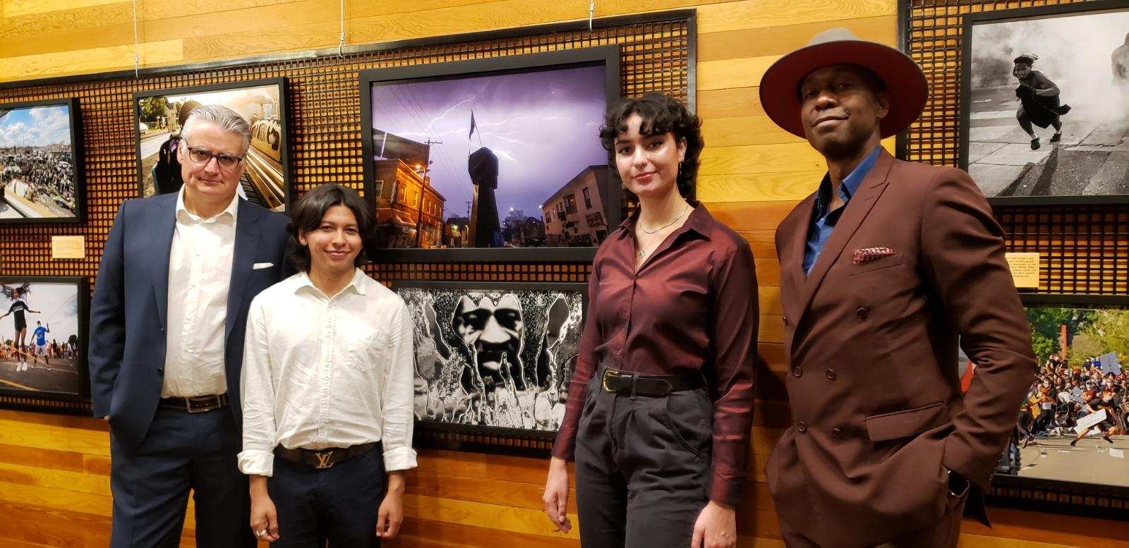 4 people stand, in fancy dress, in front of a photo gallery wall.