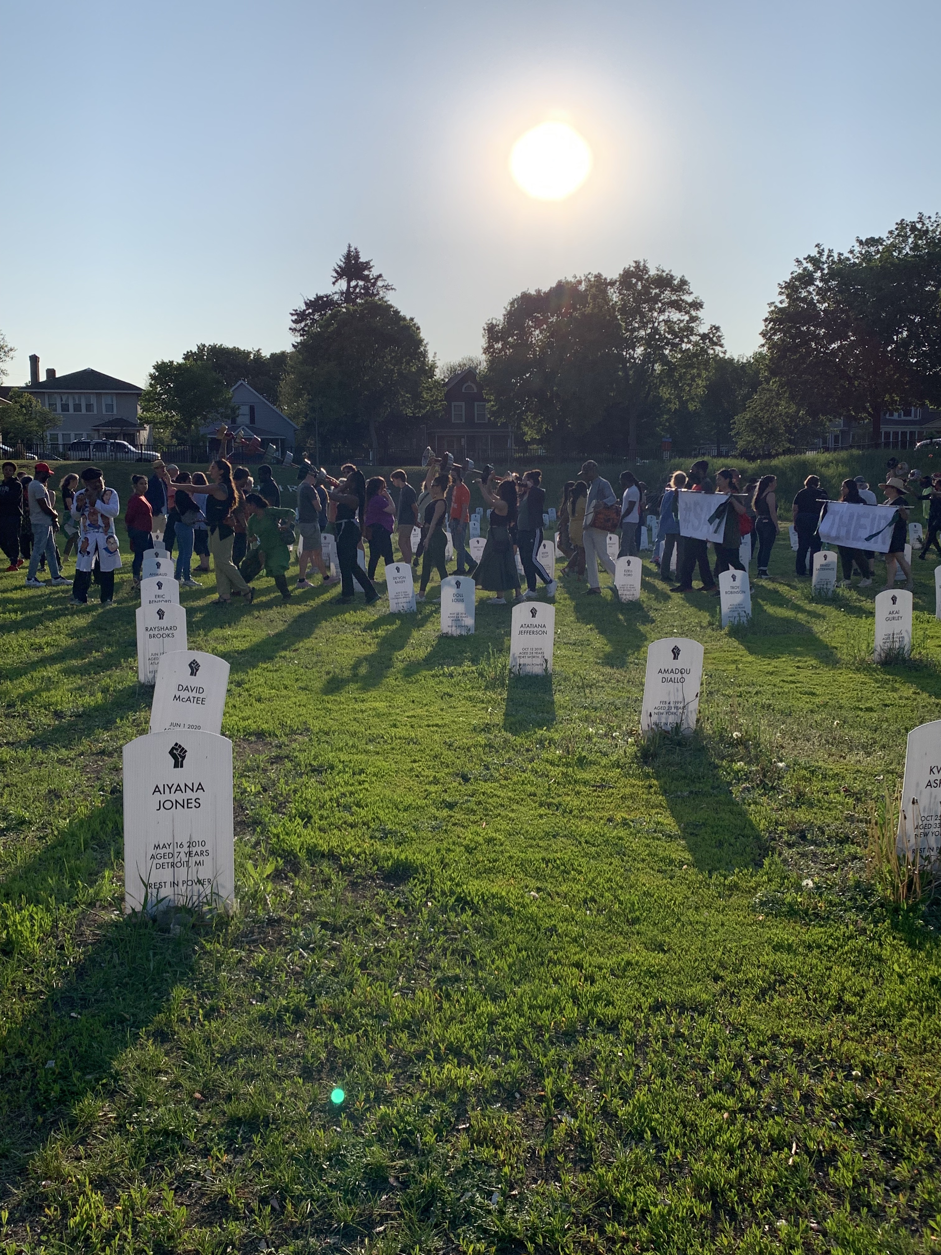 a large group of mourners processes through a field of white headstones.