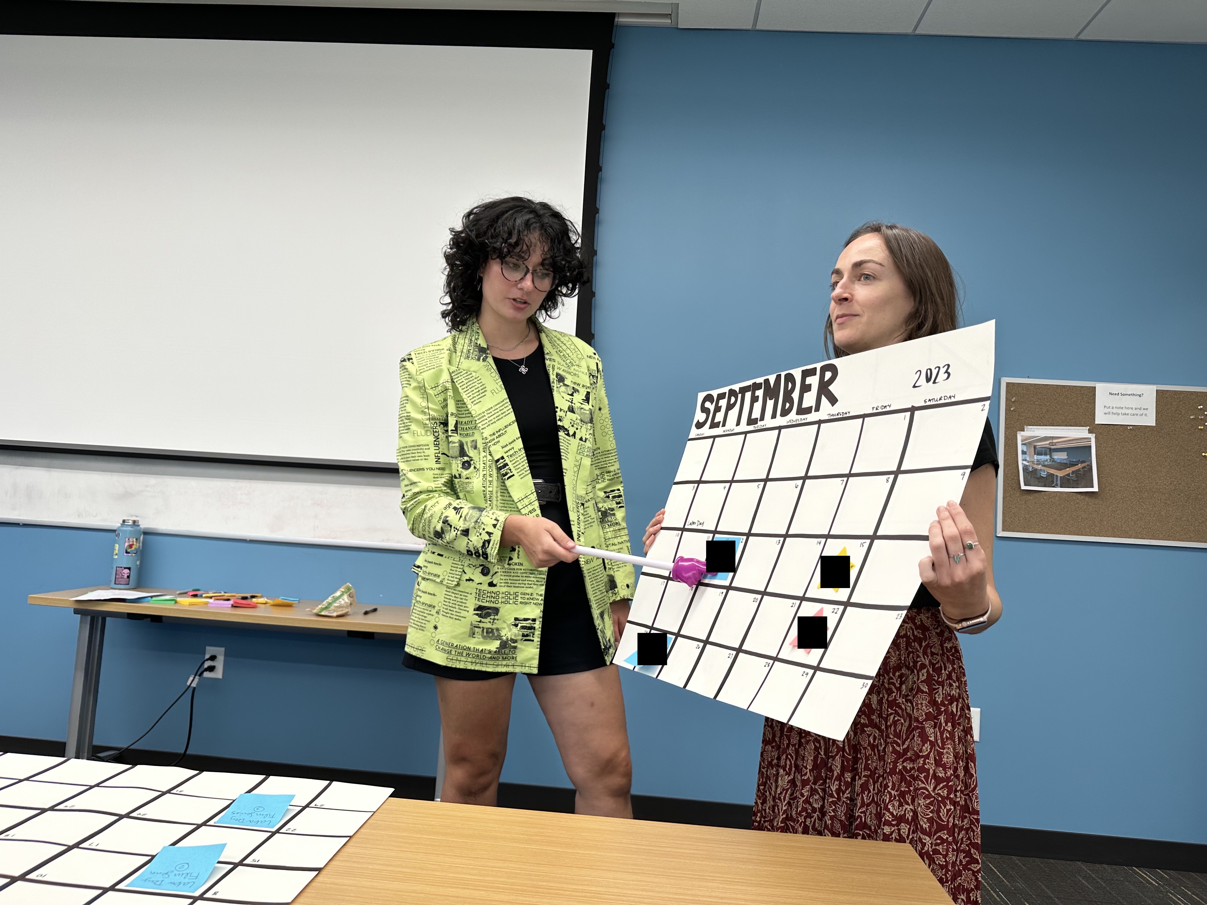 Two women hold up and point to a large calendar page.