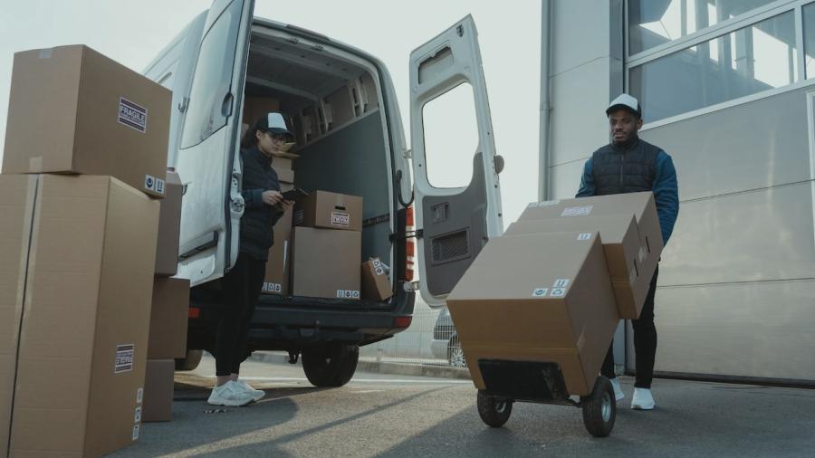 Two workers move cardboard boxes out of a sprinter van, one uses a dolly