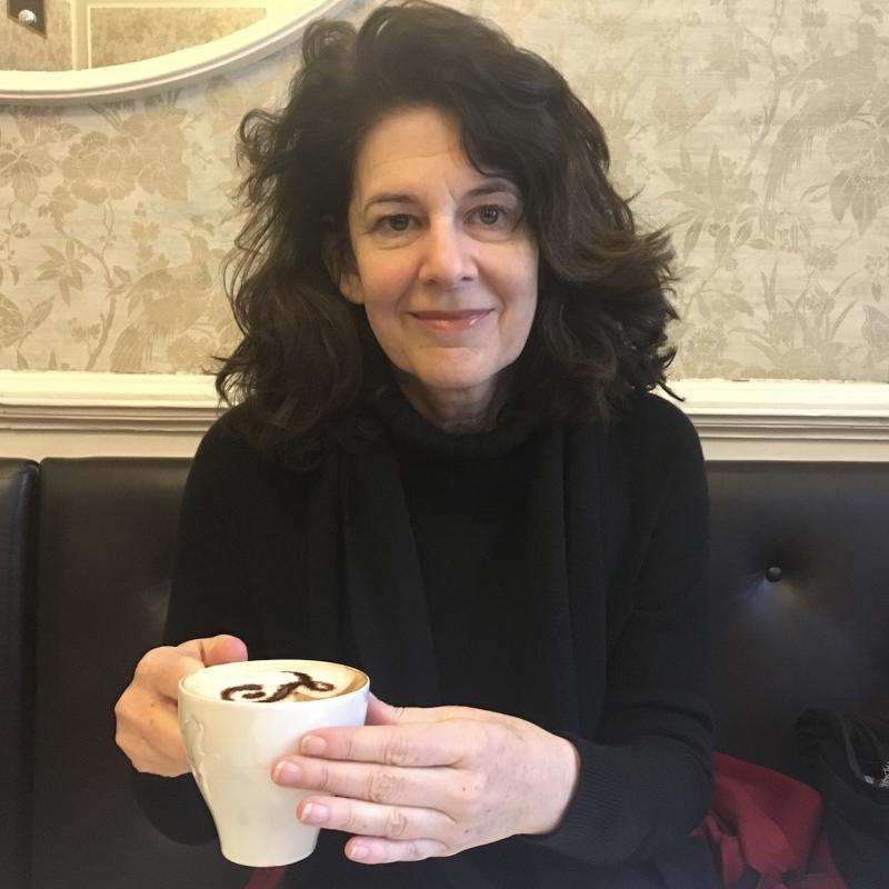 Carolyn Forbes, sitting and holding a coffee.