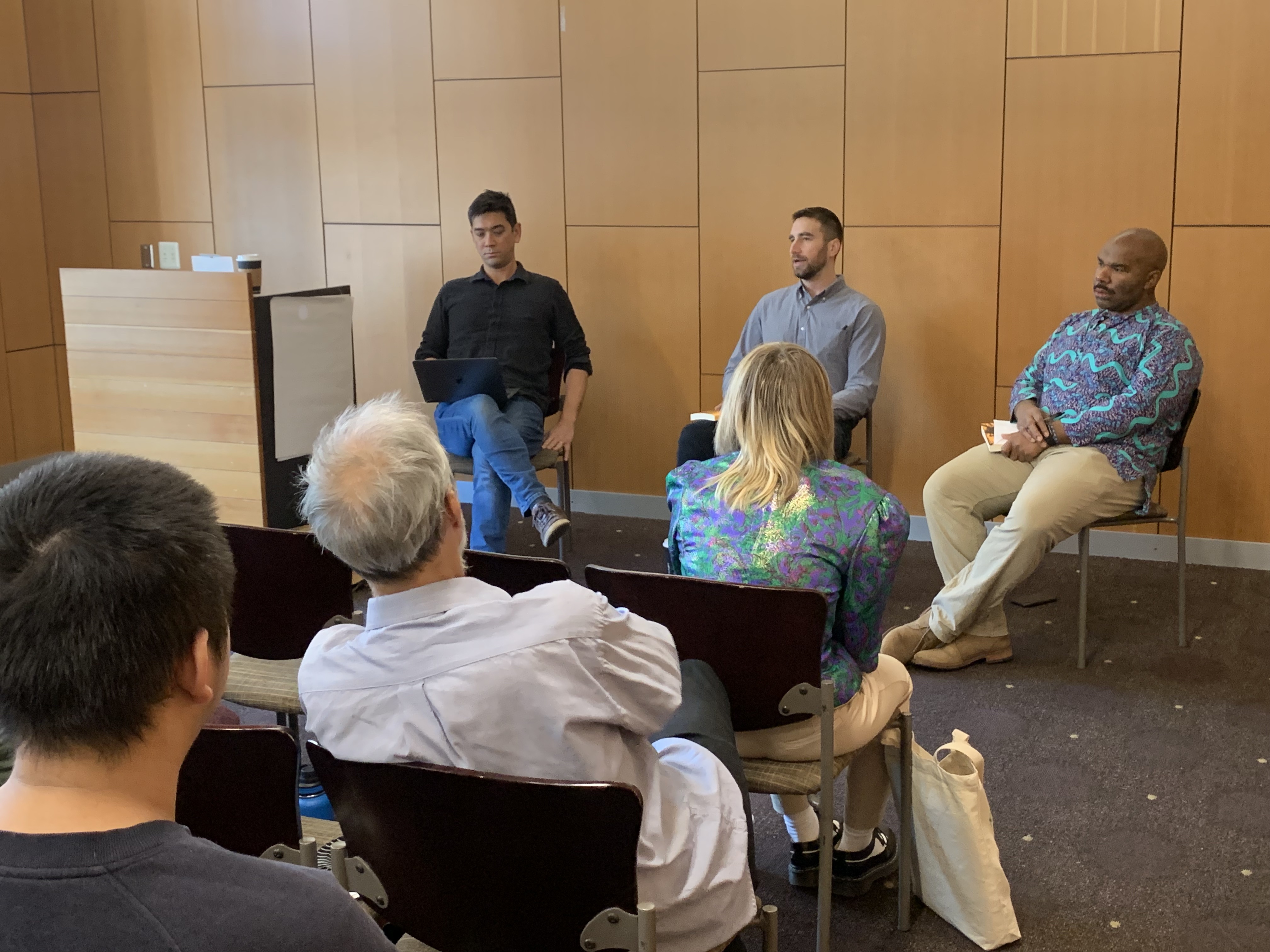 Three men sit at the front of a conference room in front of a crowd.