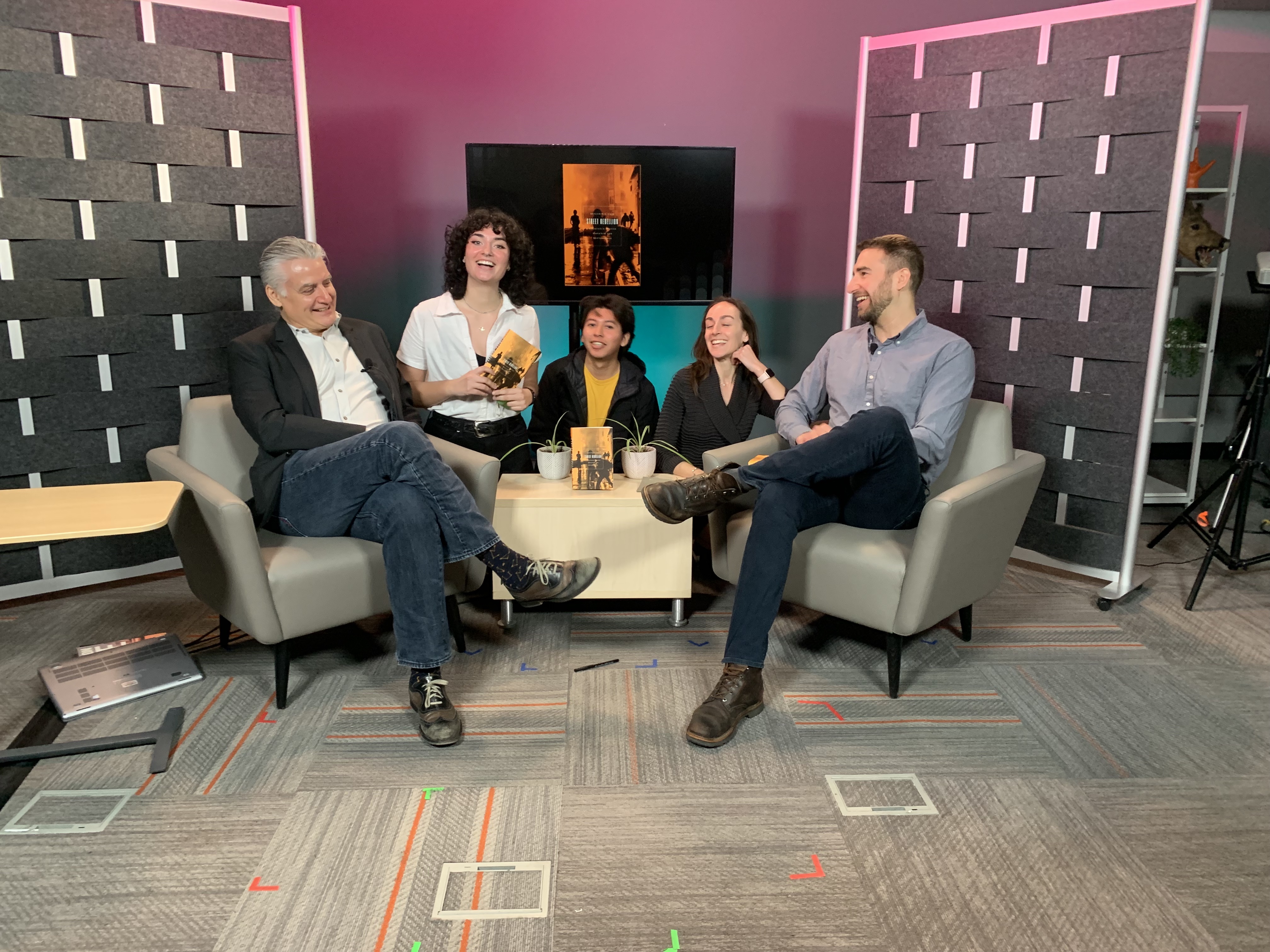 five people laugh, seated on a film set.