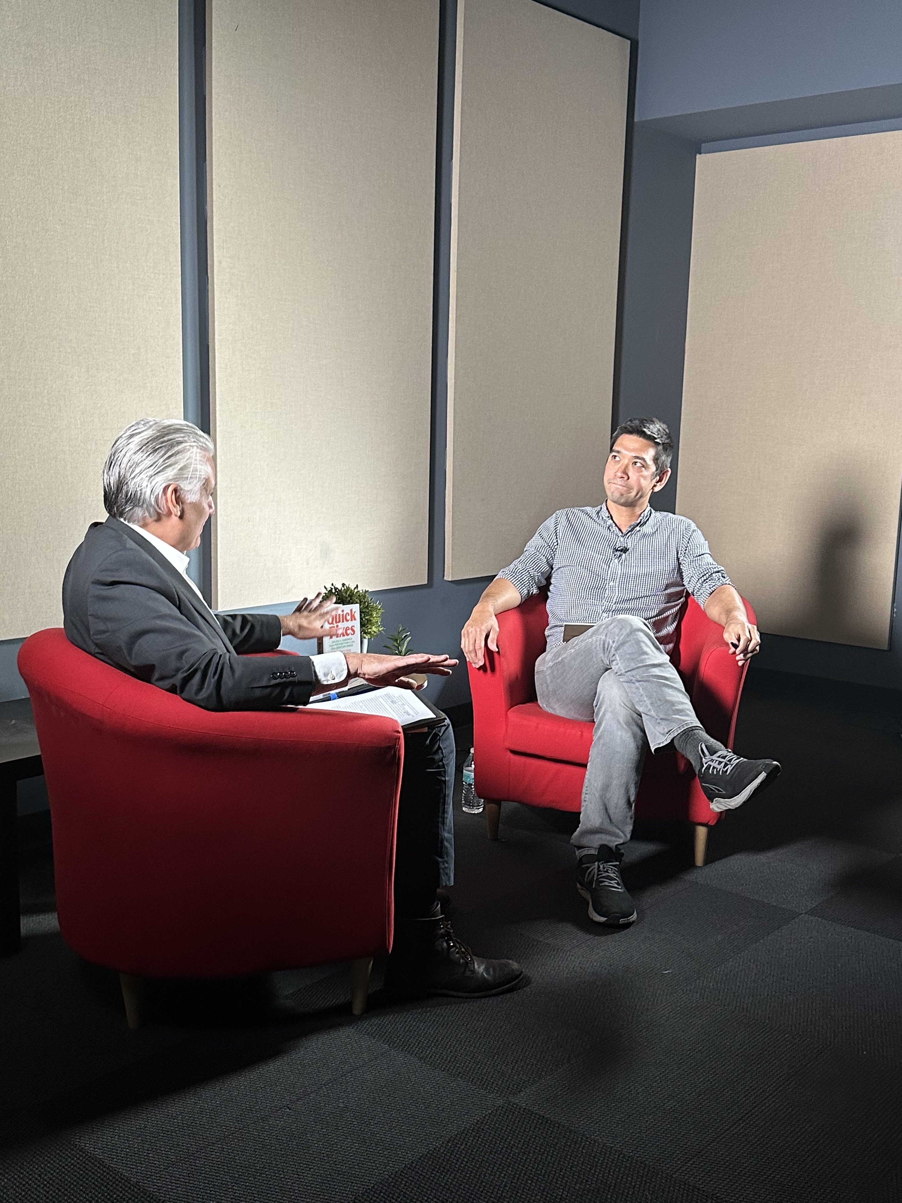 Two men sitting in red chairs speak to one another in a studio space.